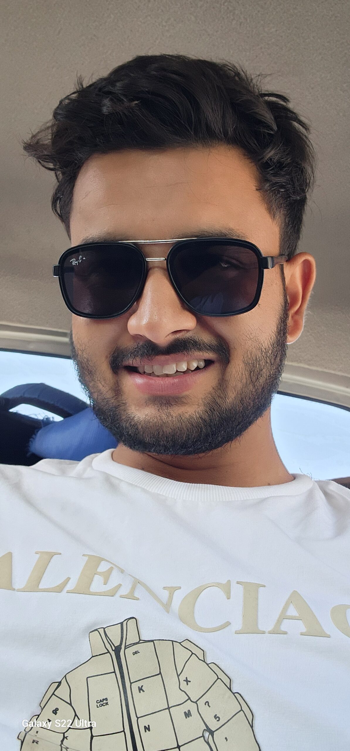 A young man with sunglasses and a beard, smiling while sitting in a vehicle. He is wearing a white t-shirt with 'Balenciaga' written on it.